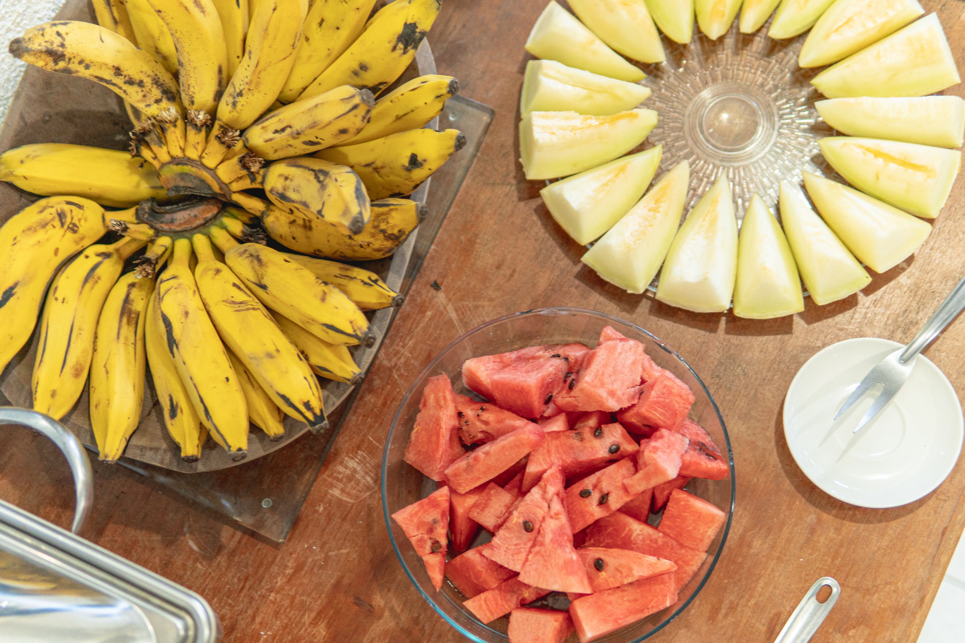 Une table sur laquelle se trouvent un plateau en métal rempli de bananes, un bol de morceaux de pastèque et une assiette en verre avec des fruits coupés en tranches.