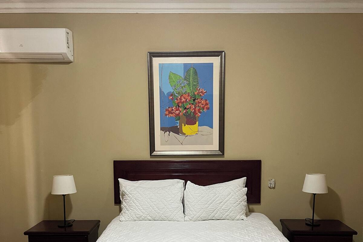 A hotel room with a bed, two bedside tables with lamps, a framed painting on the wall, and an air conditioning unit.
