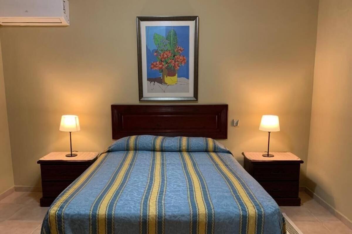 A bedroom with a bed, two bedside tables with lamps, a framed picture on the wall, and a vent on the floor.