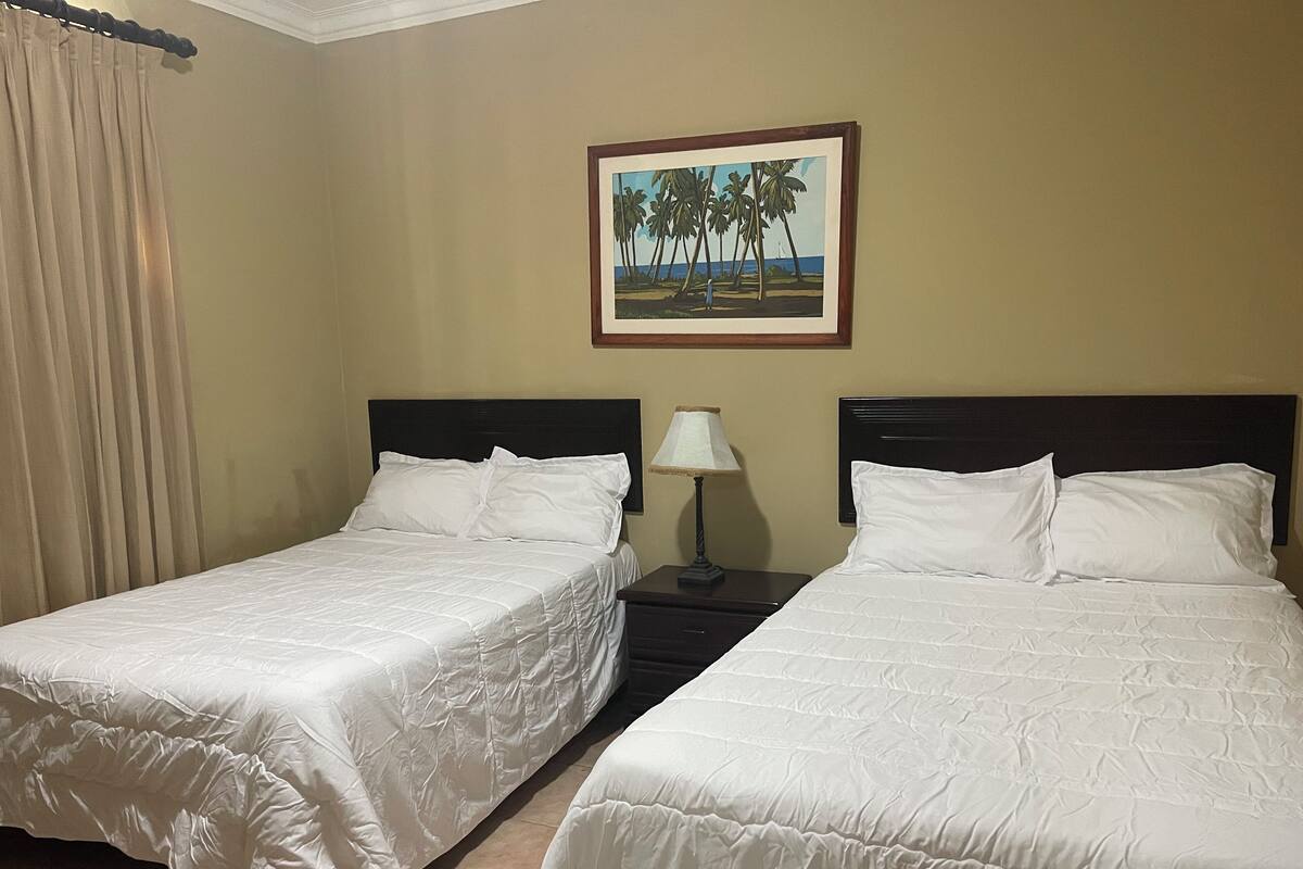A hotel room with two single beds, a nightstand with a lamp, and a painting of palm trees on the wall.