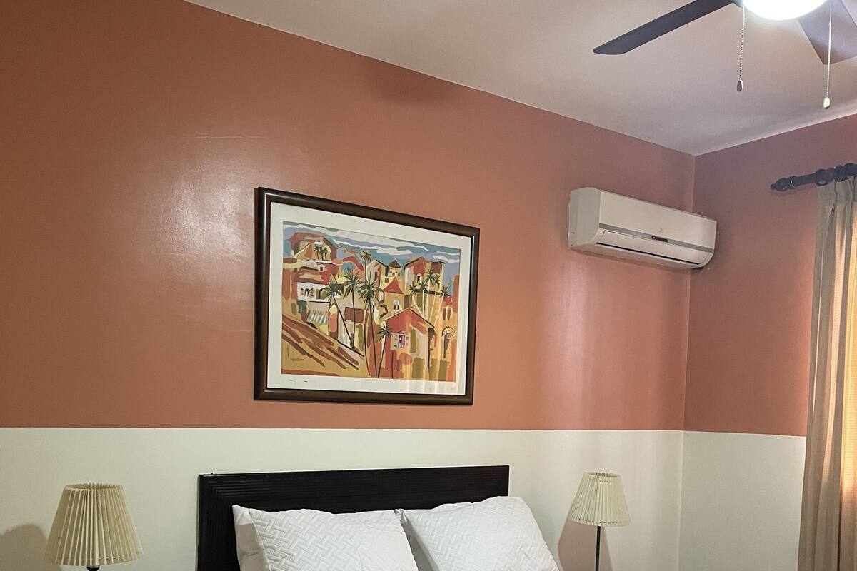 A hotel room with a bed, two bedside lamps, a ceiling fan, an air conditioning unit, and a framed painting on the wall.