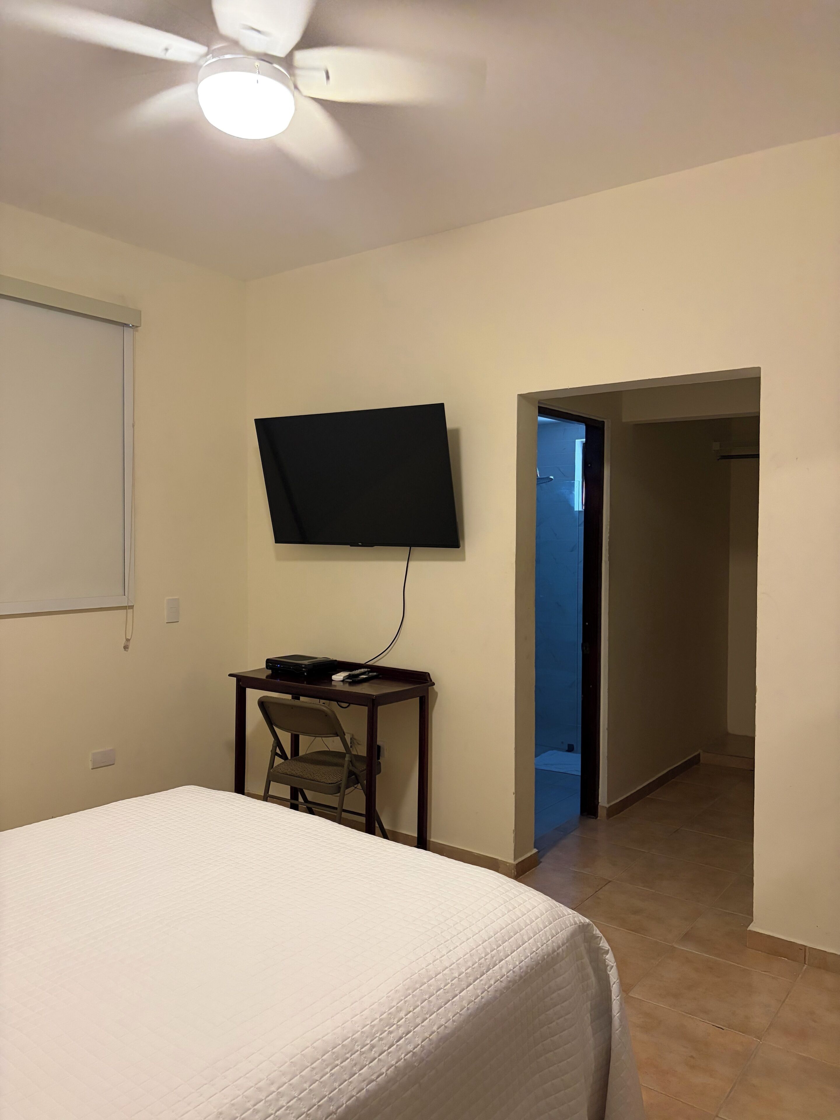 Quarto de hotel com cama, escrivaninha, cadeira, televisão de tela plana e ventilador de teto.