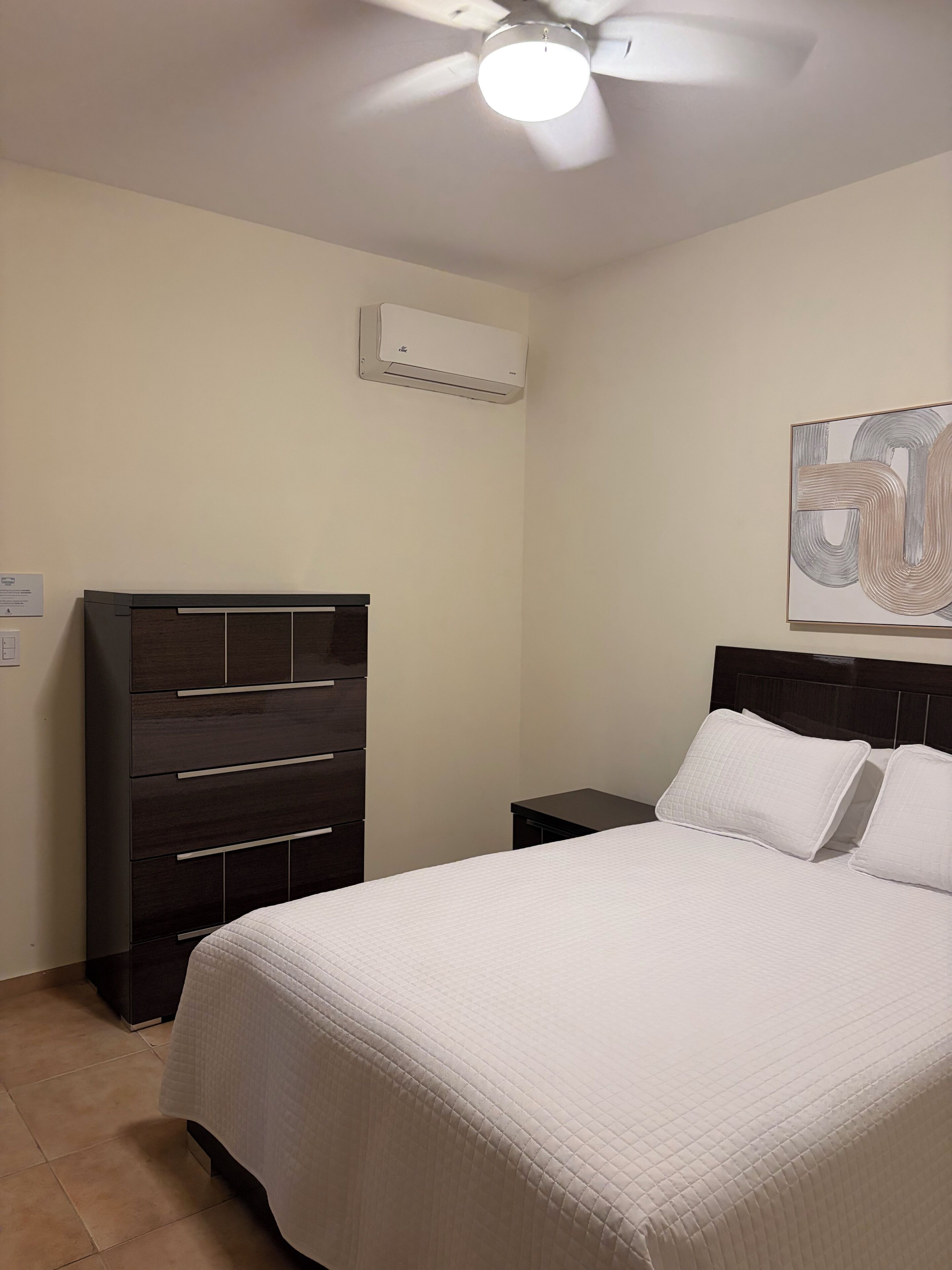 Un dormitorio con cama, cómoda, mesita de noche, aire acondicionado y ventilador de techo.