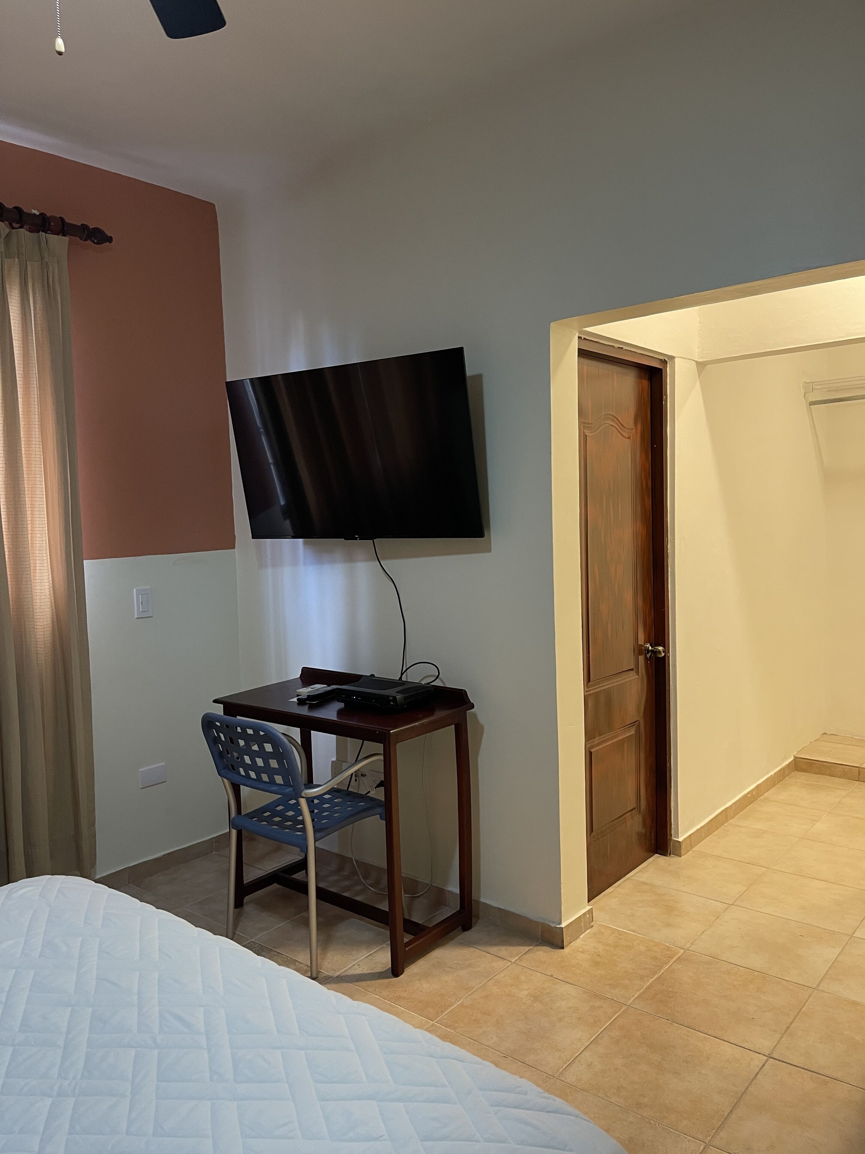 A bedroom with a bed, a desk with a chair, a television mounted on the wall, and a door leading to another room.