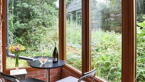 Een hoekkamer met een bakstenen muur, een raam en een tafel met stoelen. Op de tafel staan een fles wijn en glazen.