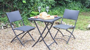 Twee zwarte metalen stoelen en een tafel met een houten plank, een brood, een bord met aardbeien en een glas water.