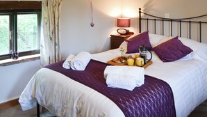 Een net opgemaakt bed met een paars sprei, witte lakens en twee opgerolde handdoeken. Er staat een dienblad met ontbijtartikelen, waaronder een koffiezetapparaat, een mok en een bord met gebak. Een bedlampje en een fotolijst zijn ook zichtbaar.