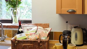 Een keukentafel met een rieten mand vol snacks, twee gele mokken, een waterkoker en een fles wijn.