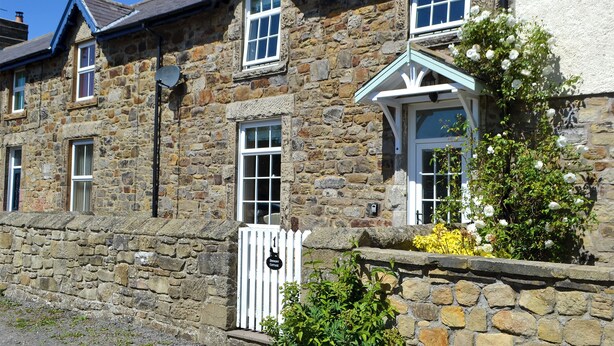 A stone building with multiple windows and a white door, surrounded by greenery and a white picket fence. - 3 bedroom accommodation in Shilbottle (Shilbottle)