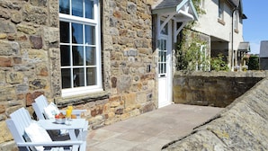 A stone building with a white door and window, outdoor seating with a table and chairs, and a stone wall. - 3 bedroom accommodation in Shilbottle (Shilbottle)