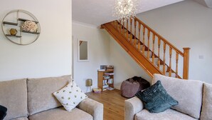 Una sala de estar con dos sofás color beige, una escalera de madera, una araña y una estantería con libros y una cesta.