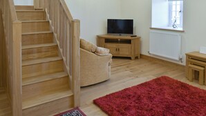 A living room with a wooden staircase, a television on a wooden stand, a window with a view, and a red rug.