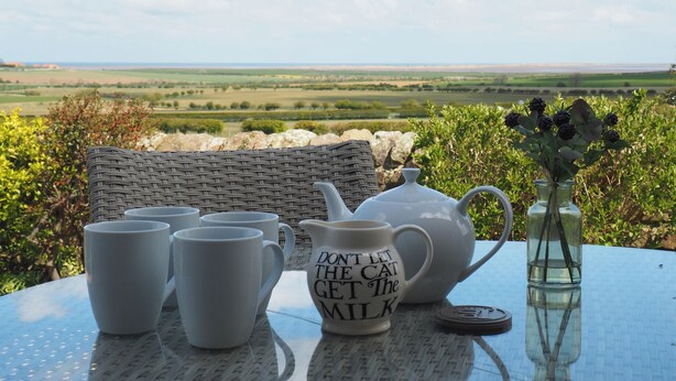 Una mesa con un juego de té y una taza con un mensaje, frente a un paisaje campestre.