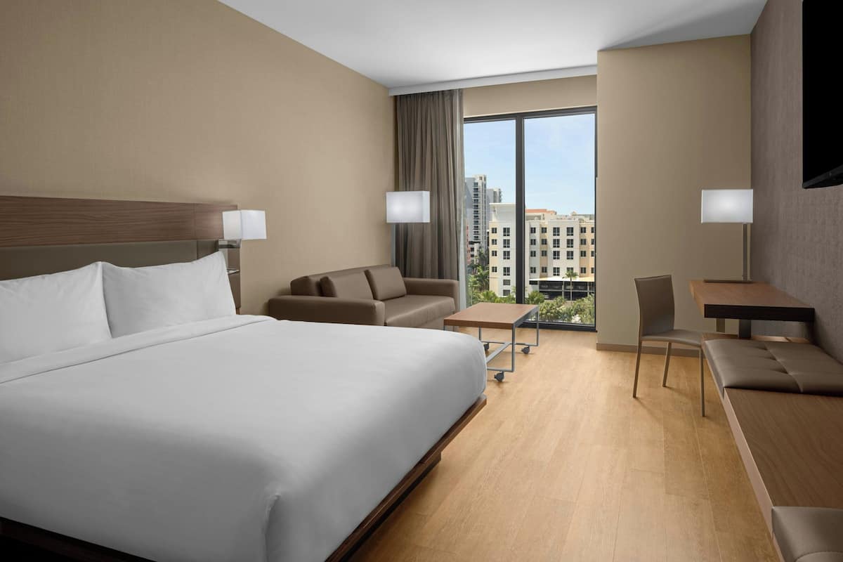 Habitación de hotel con una cama grande, un sofá, una mesita, una silla y un escritorio. Cuenta con un ventanal con vistas a edificios.
