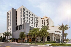 Un hotel moderno con el logotipo destacado de AC, varios pisos y un área exterior bien cuidada con palmeras.