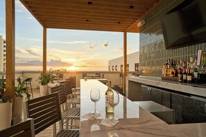 Un área de bar al aire libre con vistas a la puesta de sol, arreglos de asientos y una variedad de botellas.