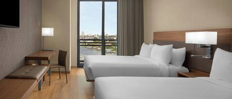 A hotel room with two beds, a desk, a chair, a TV, and a view of the city.
