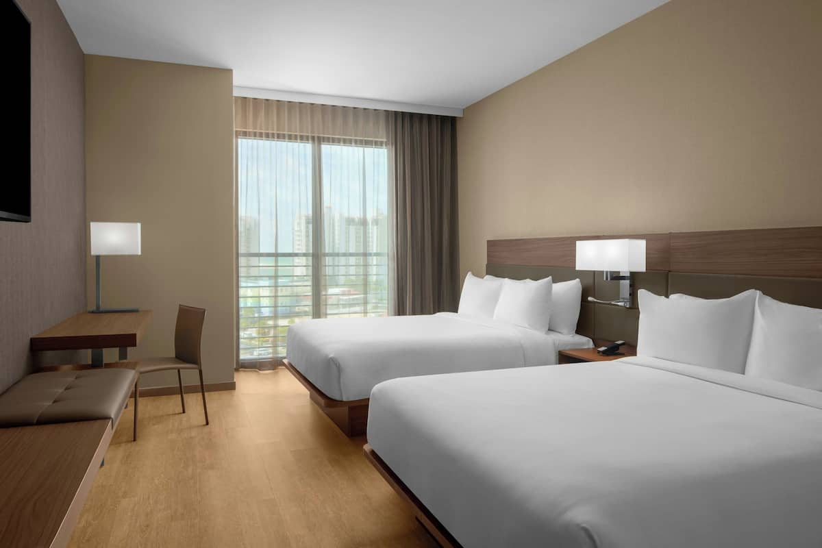 Habitación de hotel con dos camas, un escritorio, una silla y un ventanal con cortinas.