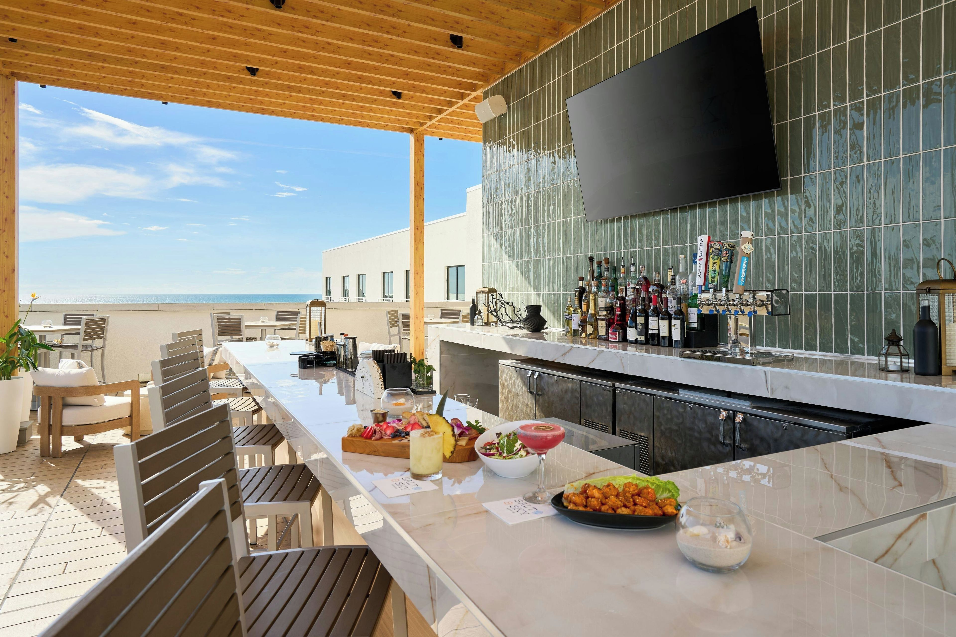 An outdoor bar area with a long counter, bar stools, and a variety of bottles and glasses.
