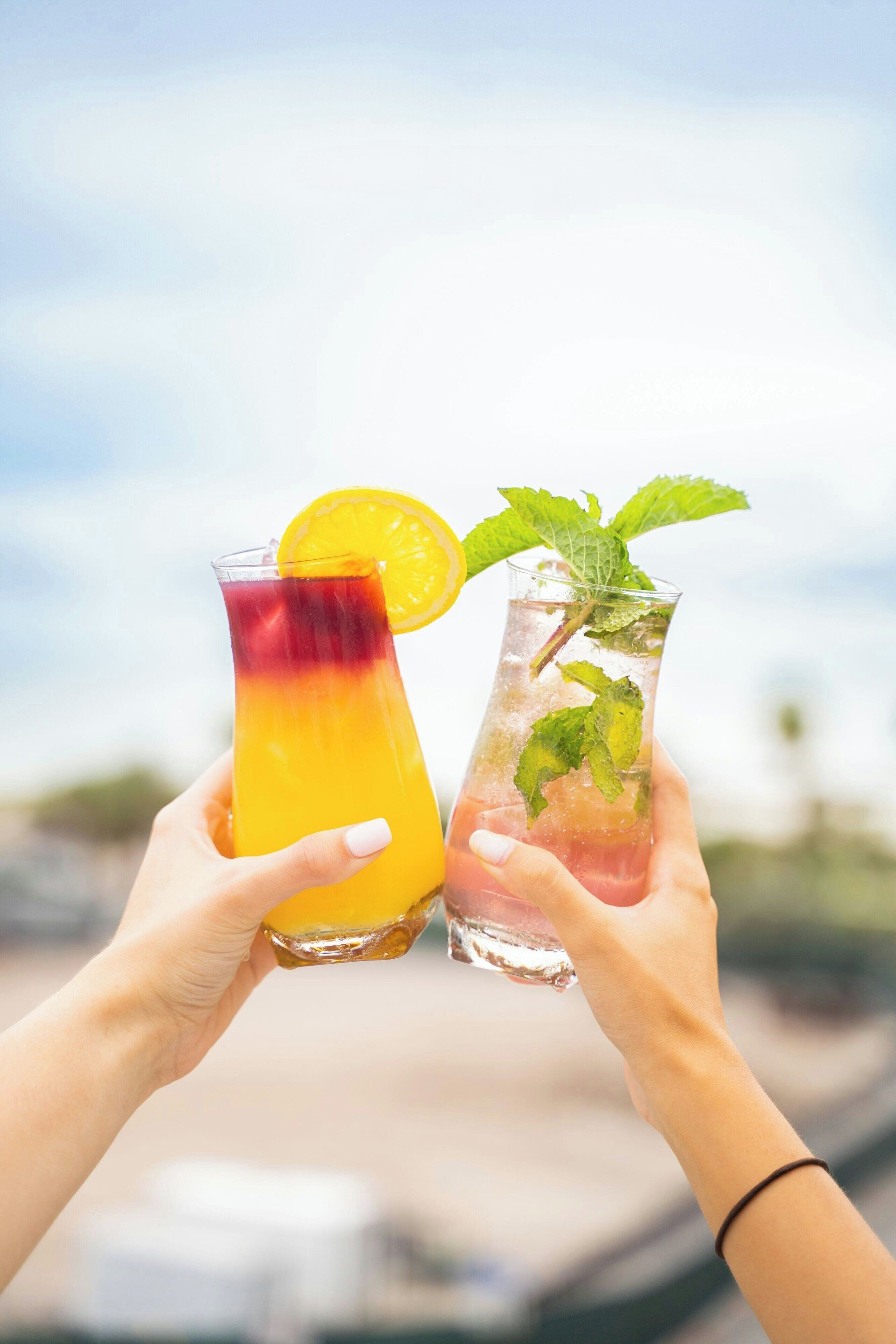 Dos manos sostienen dos vasos de bebidas heladas, uno con un degradado de naranja a rojo y el otro con hojas de menta y una rodaja de lima.