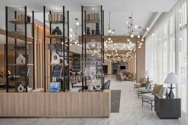 A modern hotel lobby with a reception desk, seating area, and a large window providing natural light.