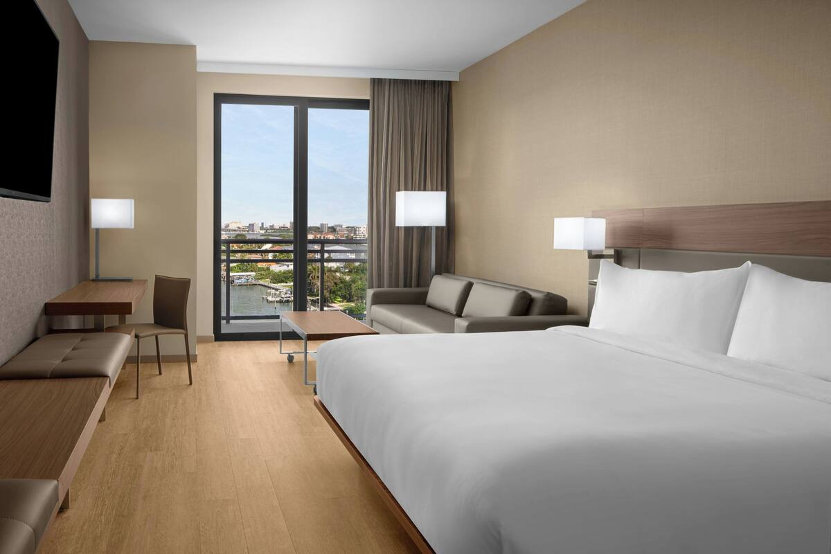Habitación de hotel con una cama grande, un sofá, un escritorio y una silla. Cuenta con un ventanal que ofrece vistas a un paisaje urbano.