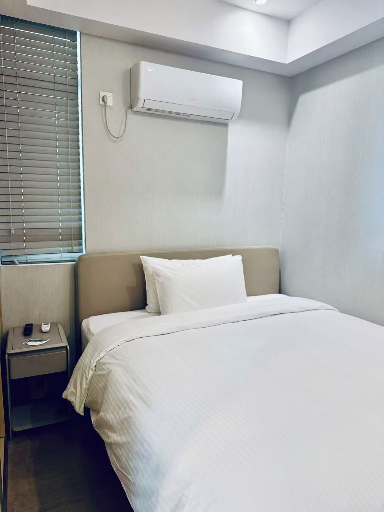 A hotel room with a large bed, a bedside table with a phone and a remote, and an air conditioning unit mounted on the wall.