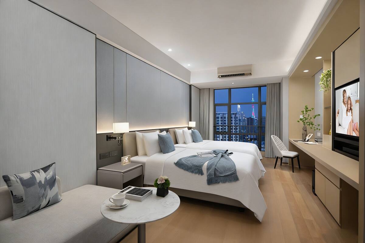 Una habitación de hotel moderna con una cama grande, un escritorio, una silla, una mesita con una taza y un libro, y un televisor.