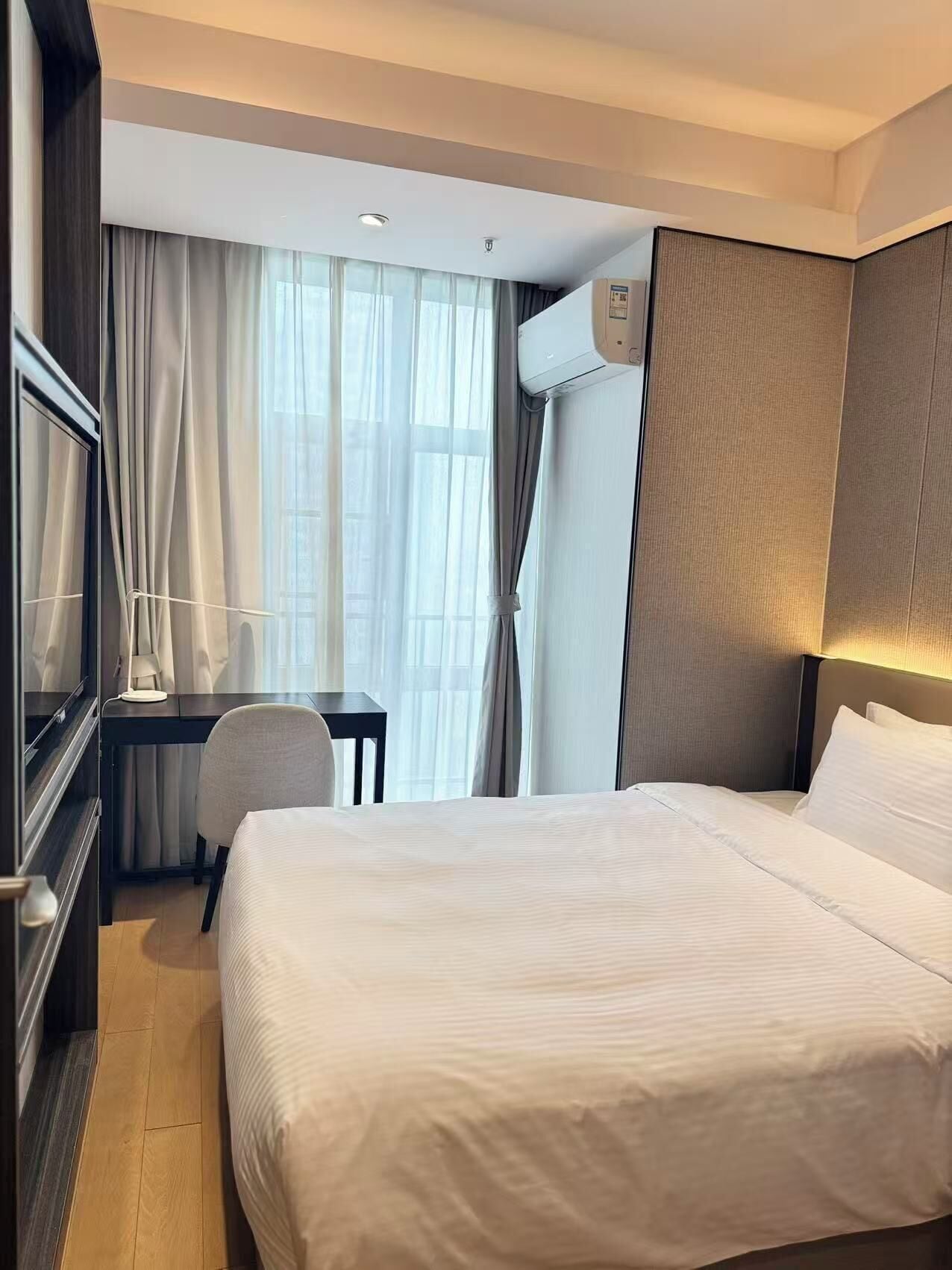 A hotel room with a bed, a desk, a chair, and an air conditioning unit.
