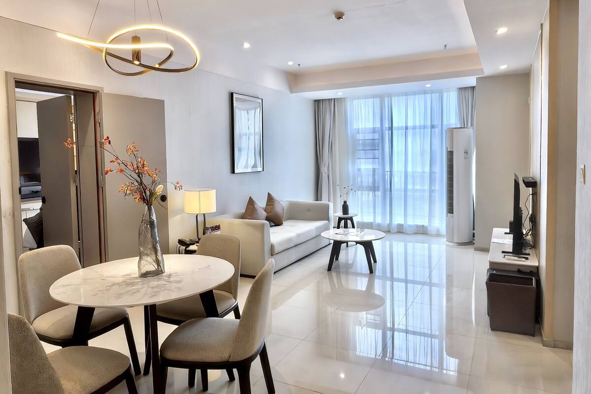 Habitación de hotel moderna con zona de comedor, sofá, mesa de centro y un amplio ventanal con cortinas.