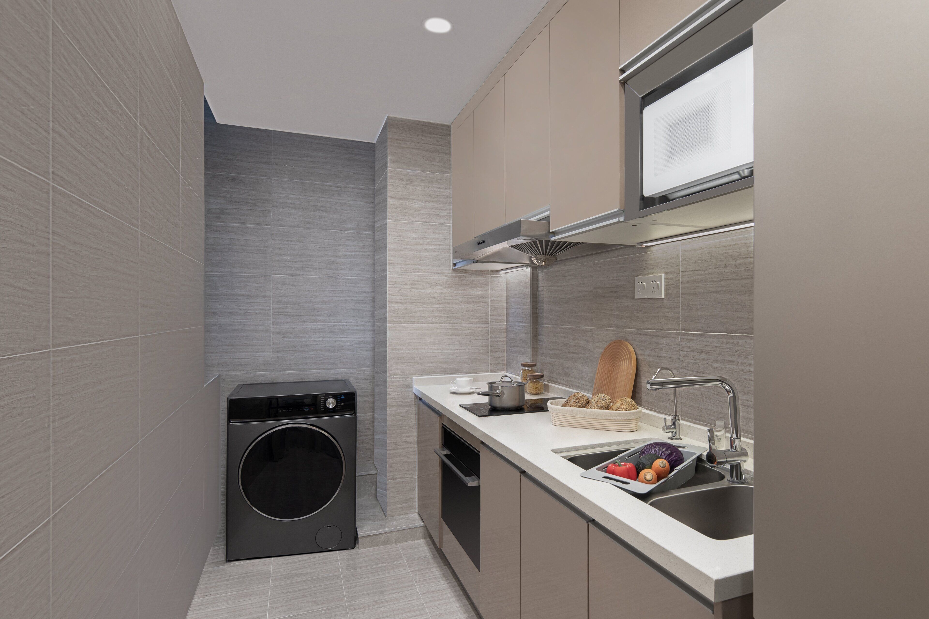 A compact kitchen with grey tiles, a built-in washing machine, a sink with a cutting board, and a window above the stove.