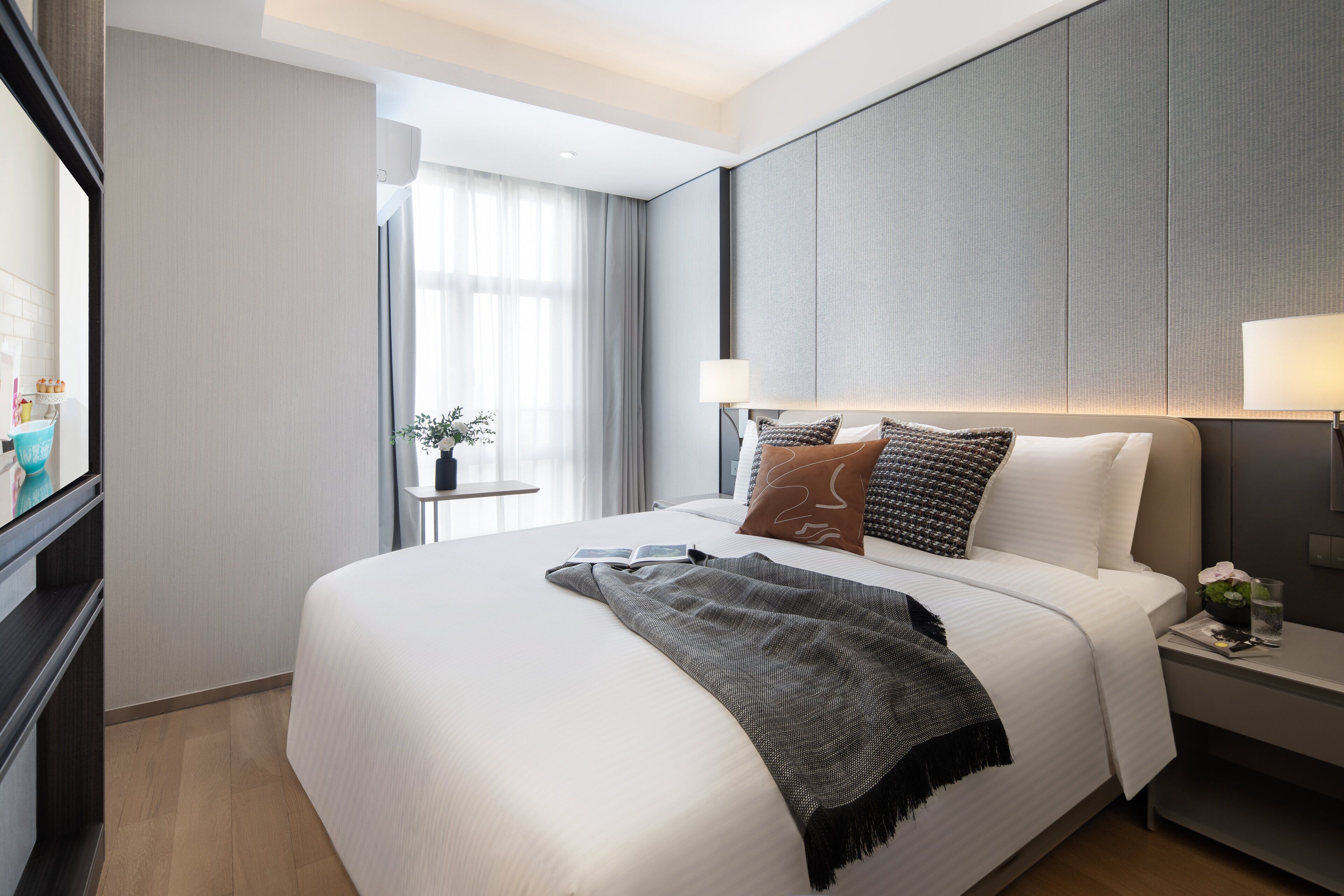A modern hotel room with a large bed, a bedside table, a nightstand, a window with curtains, and a view of the cityscape.