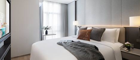 A modern hotel room with a large bed, a bedside table, a nightstand, a window with curtains, and a view of the cityscape.