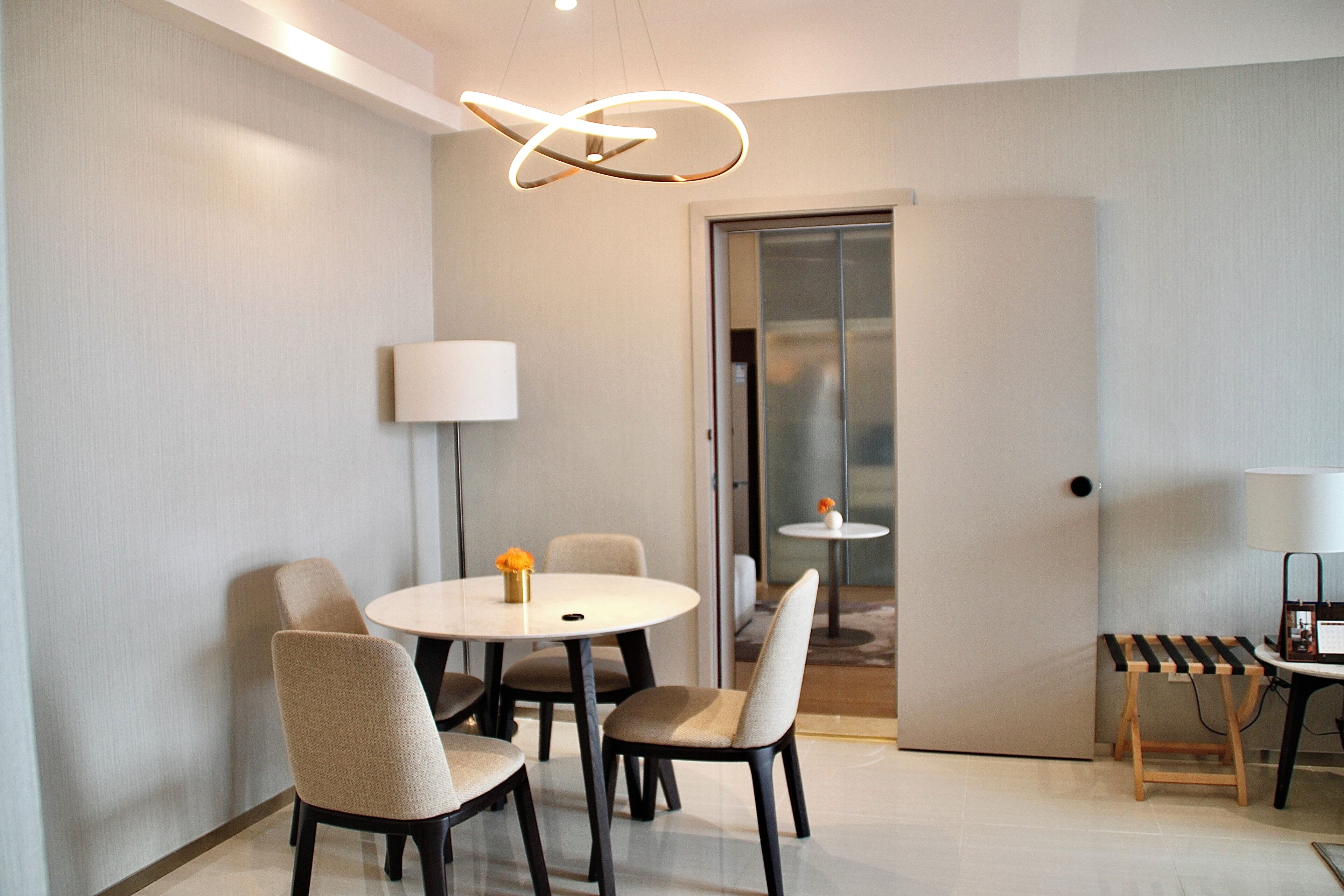 A modern dining area with a round table, four chairs, a hanging light fixture, and a small side table with a lamp.