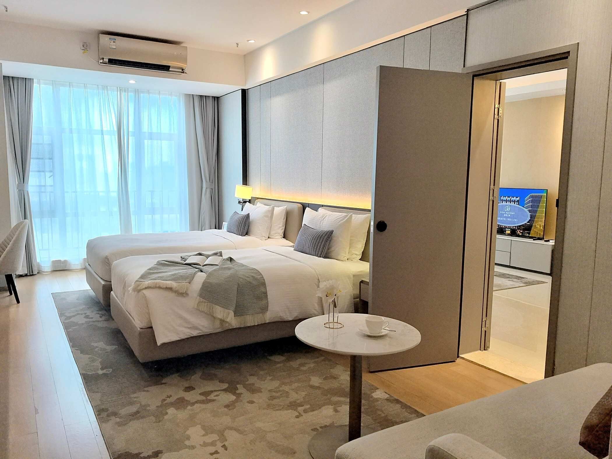 A modern hotel room with two beds, a small round table with a cup, and a view of the city through the window.