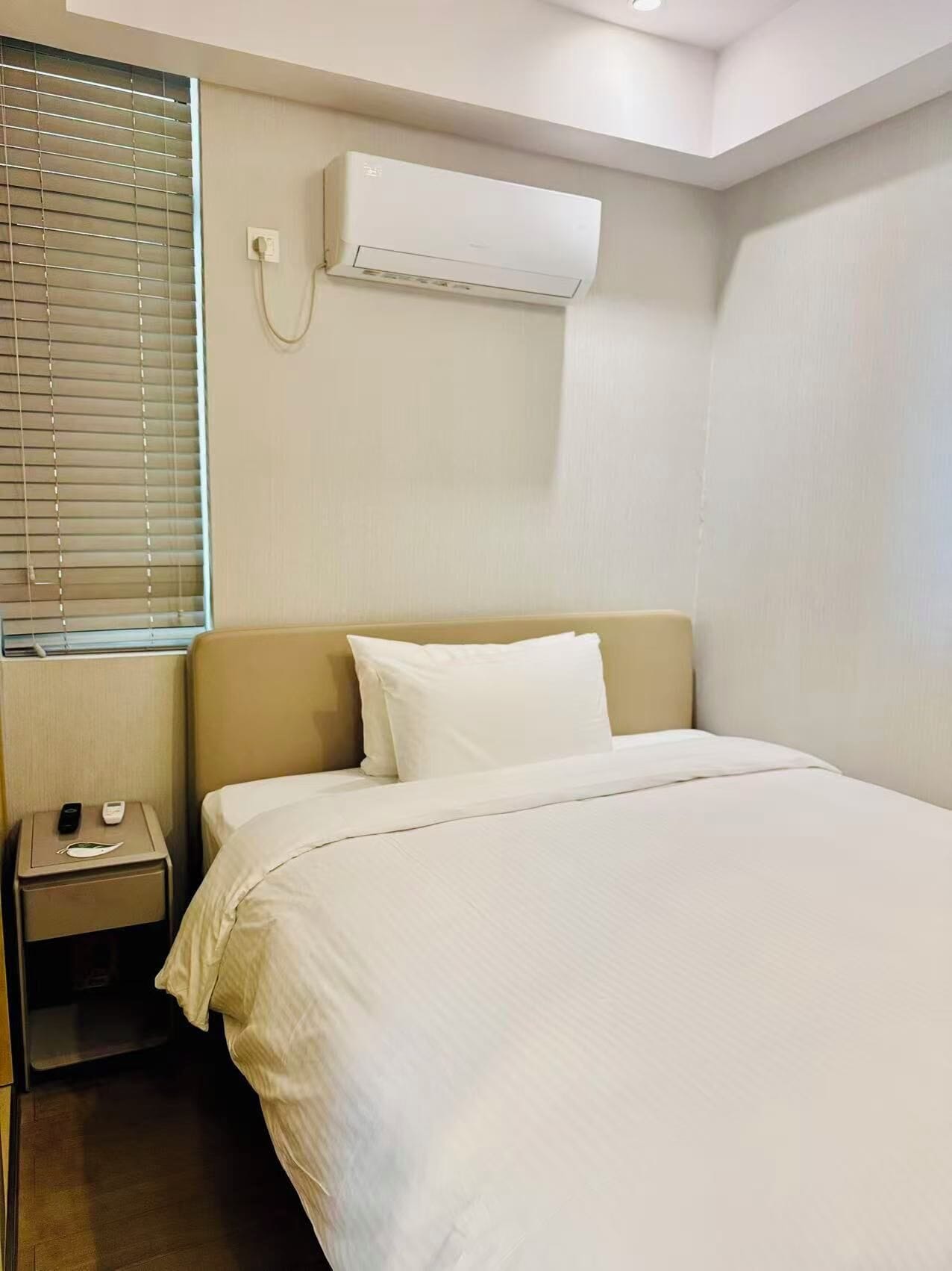 A hotel room with a bed, a bedside table with a phone and a remote, and an air conditioning unit mounted on the wall.