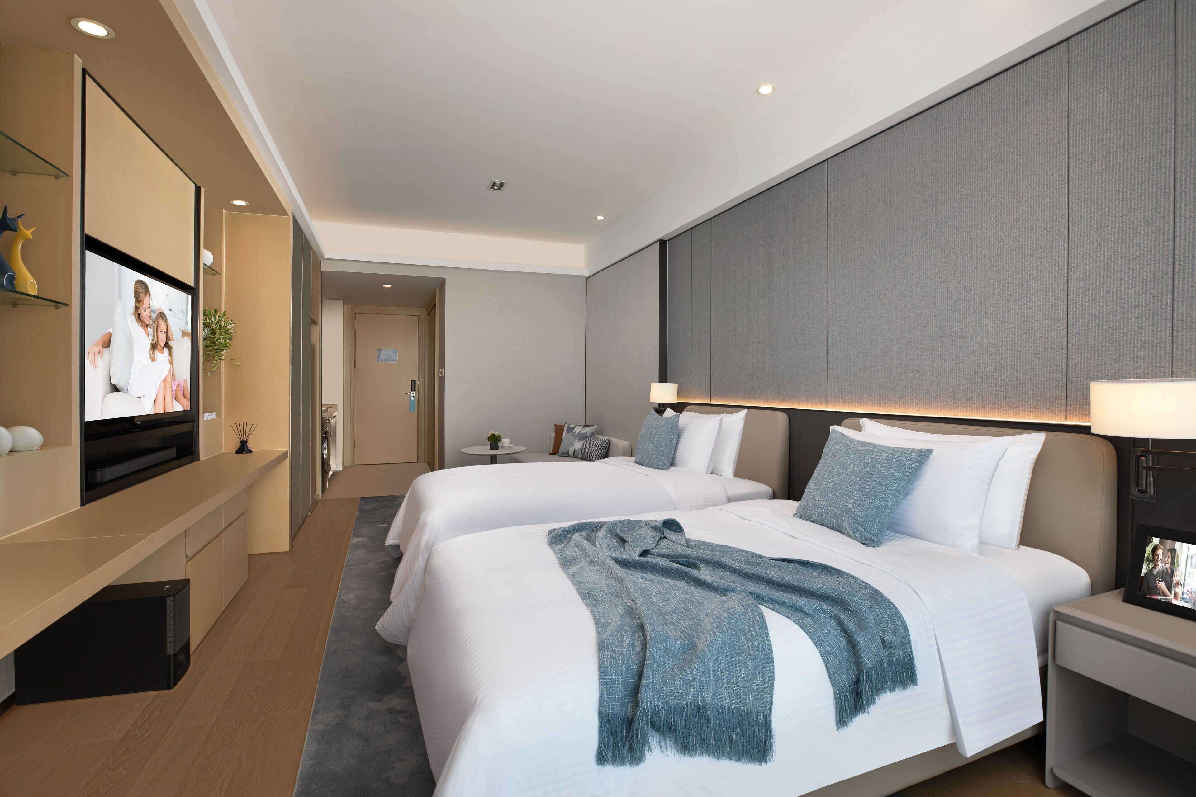 A modern hotel room with two beds, a TV on a stand, a bedside table with a lamp, and a large mirror.