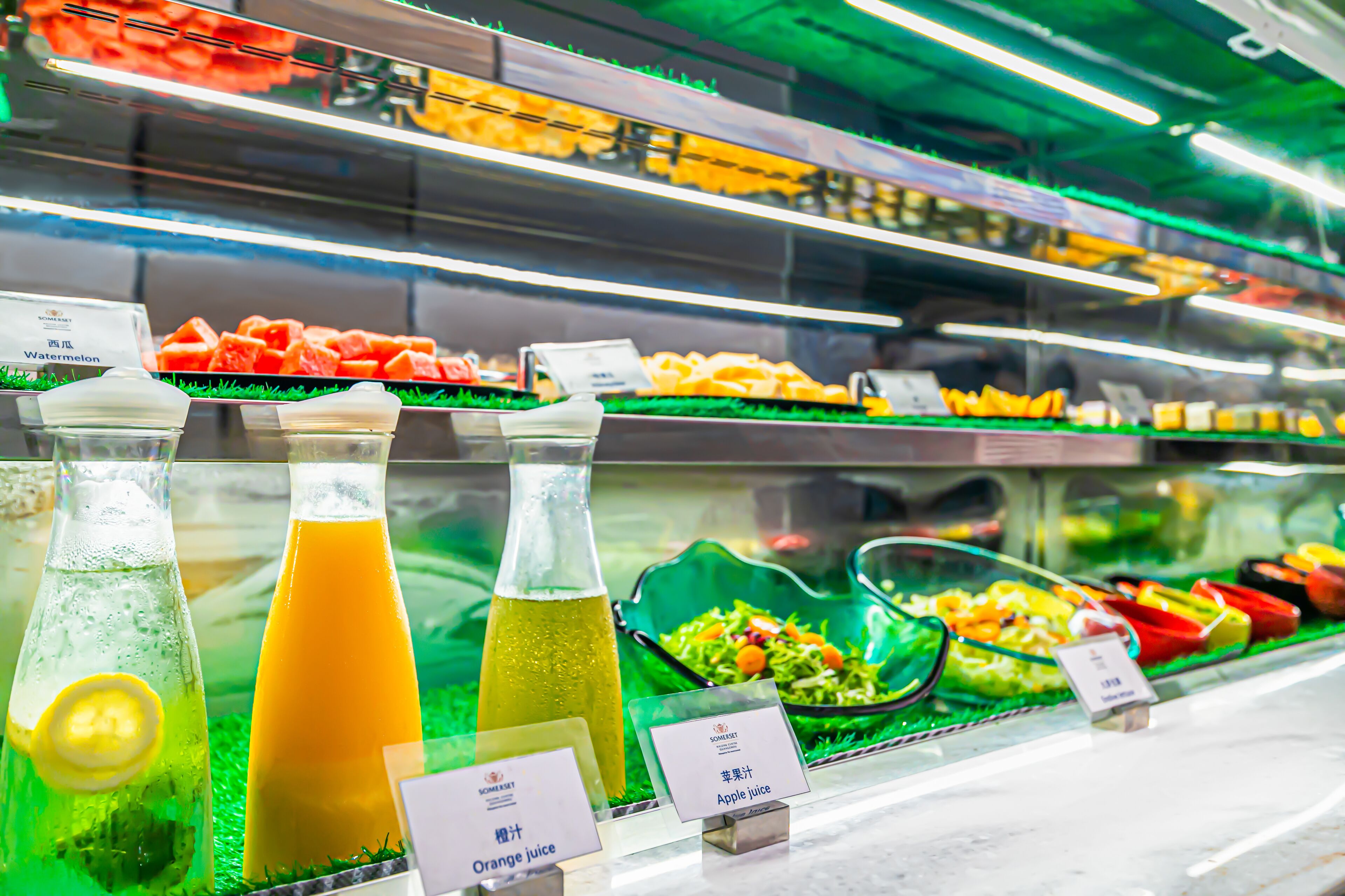 Una barra de ensaladas con diversas frutas y verduras, entre ellas sandía y zanahorias, y opciones de jugos identificadas como naranja y manzana.