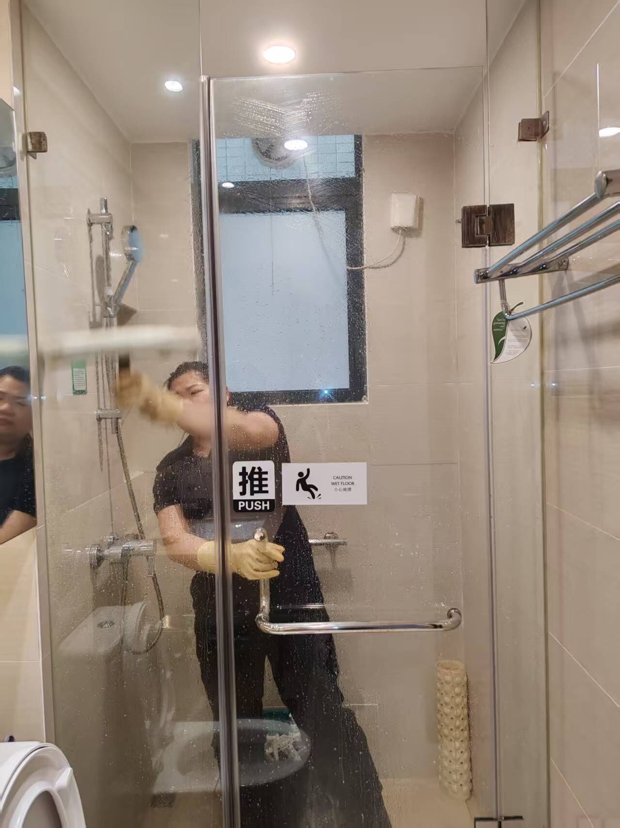 A glass-enclosed shower stall with a push bar, a caution sign for a wet floor, and a window with a view of the outside.