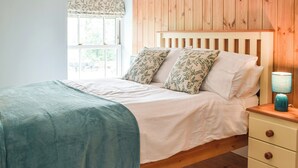 Un dormitorio con cabecera de madera, cama con sábanas blancas y dos almohadas estampadas, mesita de noche con lámpara y ventana con vistas a zonas verdes.