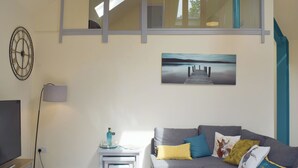 Een moderne woonkamer met een grijze bank, een flatscreen televisie, een klein wit tafeltje met een lamp en een schilderij van een pier.