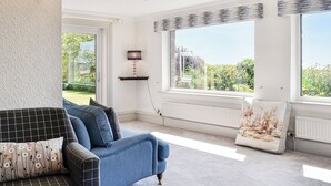 A living room with a blue sofa, a patterned armchair, and a view of greenery through large windows. - 4 bedroom accommodation in Annan (Annan)