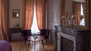 Quarto com piso de madeira, duas poltronas roxas, uma mesa redonda com um copo, lareira e um espelho grande refletindo três copos de vinho.
