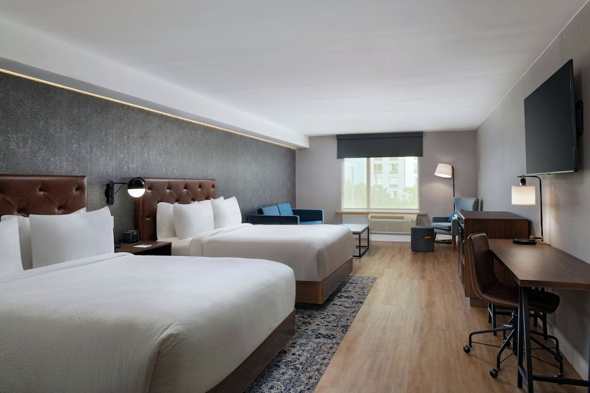 A hotel room with two beds, a desk, a chair, a TV, and a window with a view of buildings.