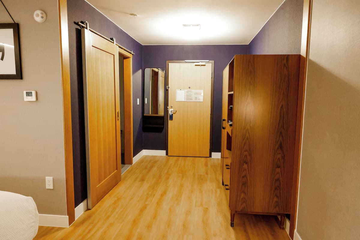 A hallway with wooden floors, a door with a mirror, and a thermostat on the wall.