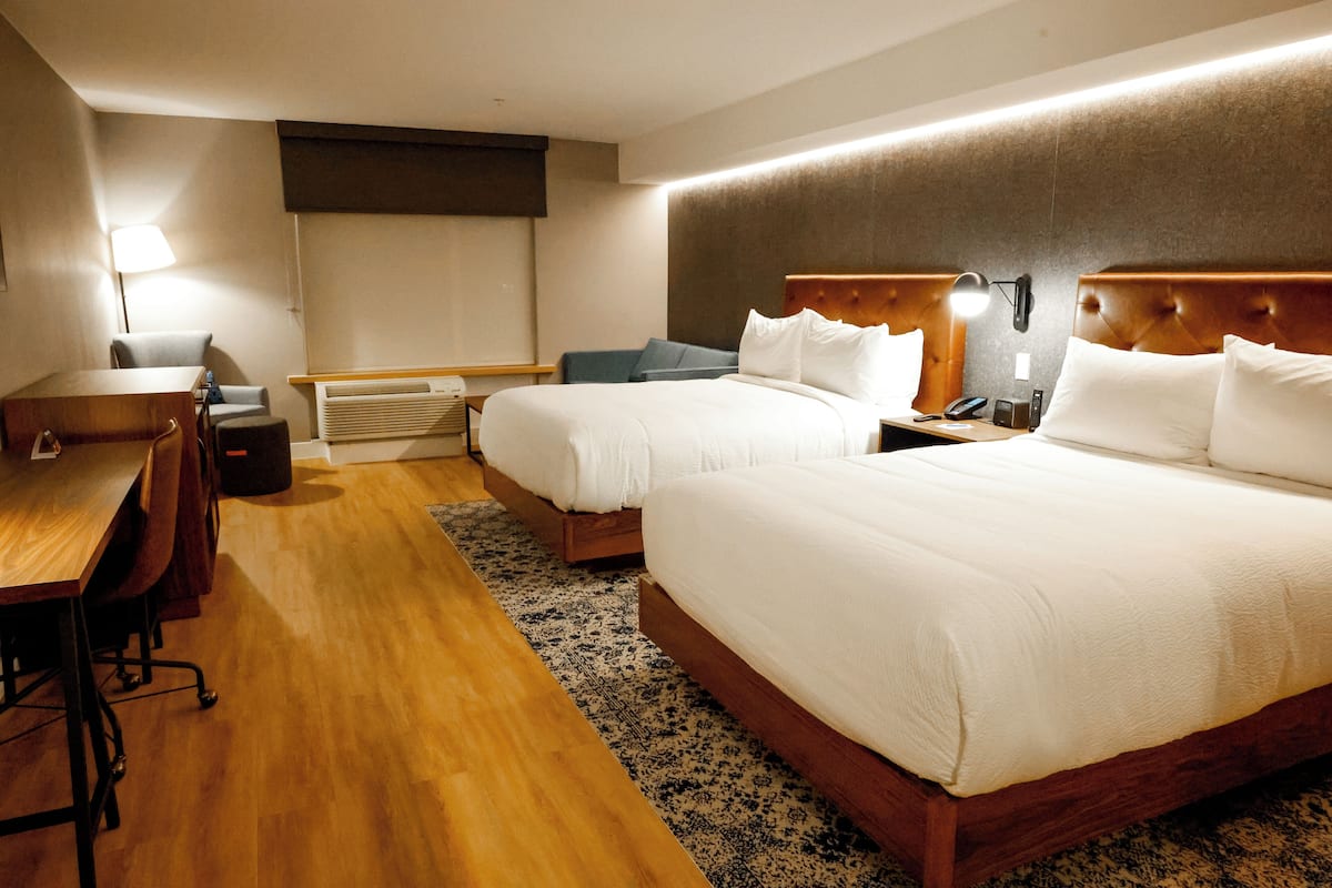 A hotel room with two beds, a desk, a chair, a lamp, and a heater.