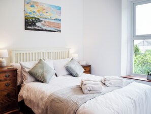 A neatly made bed with white linens and two pillows, a bedside table with a lamp, and a window with a view of greenery. - 4 bedroom accommodation in Pwll near Llanelli (Pwll near Llanelli)