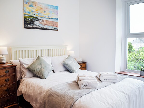 A neatly made bed with white linens and two pillows, a bedside table with a lamp, and a window with a view of greenery.