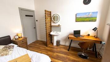 Un dormitorio con cama, mesita de noche, lámpara, escalera de madera, lavamanos, espejo, ordenador portátil, silla y un cuadro en la pared.