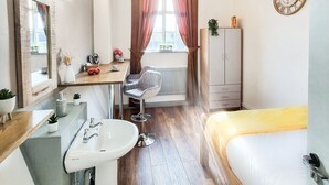A modern bedroom with a wooden floor, a bed with a yellow blanket, a bedside table, a wardrobe, a large window with curtains, a mirror, a sink, and a chair. - Cades Accommodation (Wolverhampton)